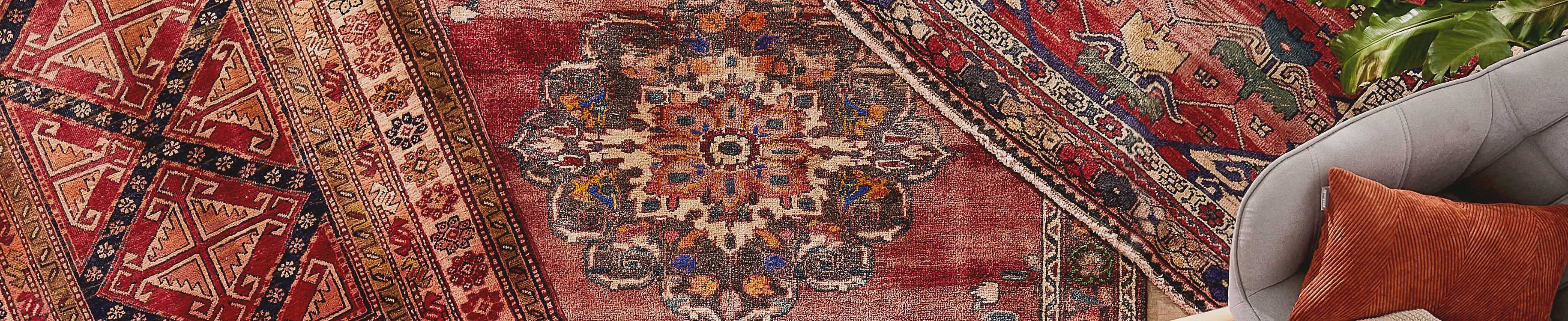 Detailaufnahme von Teppich mit Muster, daneben grauer Sessel mit orangefarbenem Kissen und Pflanze.