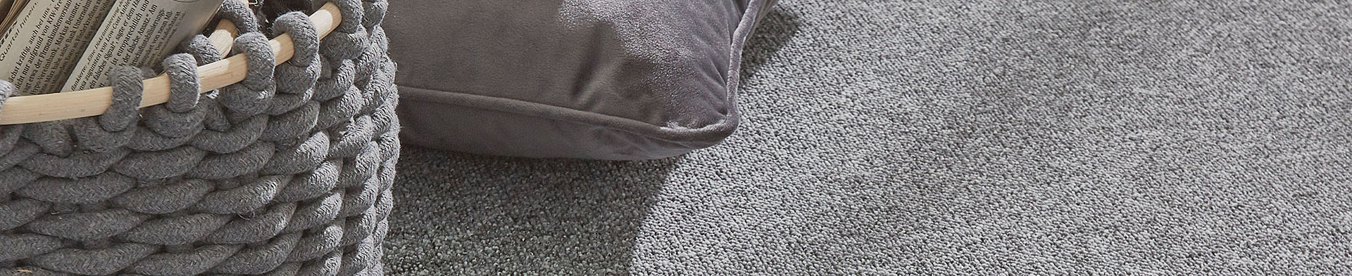 Geknüpfter Korb hält Zeitung, liegt neben einem Kissen auf grauem Teppichboden.