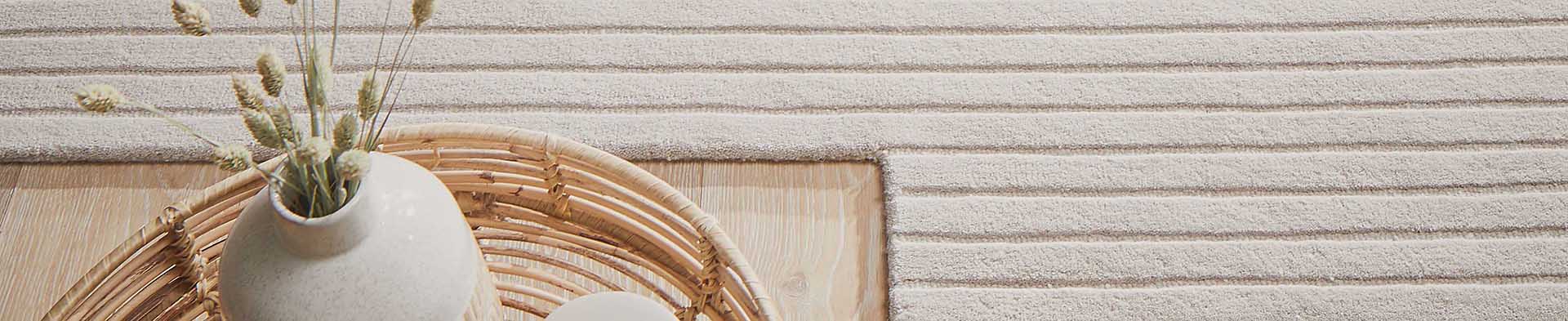 Vase mit getrockneten Pflanzen steht im Korb auf Holztisch neben gestreiftem Teppich.