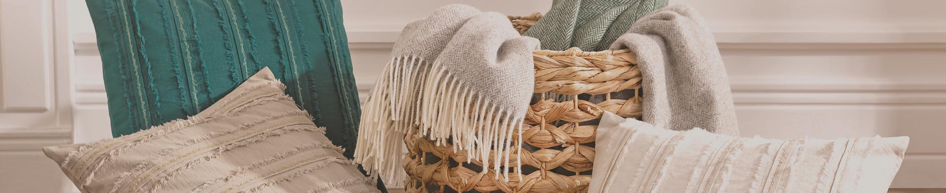 Kissen und Decken liegen locker in einem geflochtenen Korb vor einer hellen Wand mit Zierleisten.