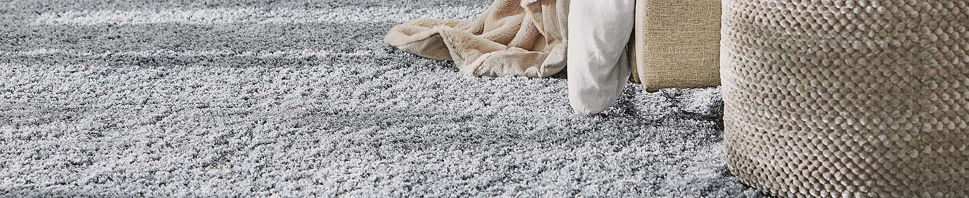 Grauer Teppich liegt unter einer Couch mit hellen Kissen und einer Decke im Wohnzimmer.