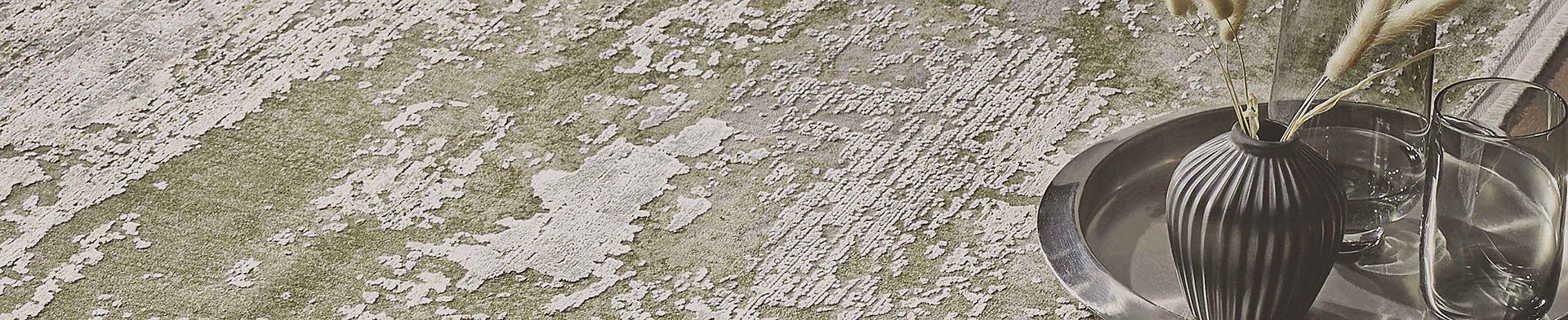 Vase mit Trockenblumen steht auf rundem Tablett neben Gläsern auf dekoriertem Teppich.