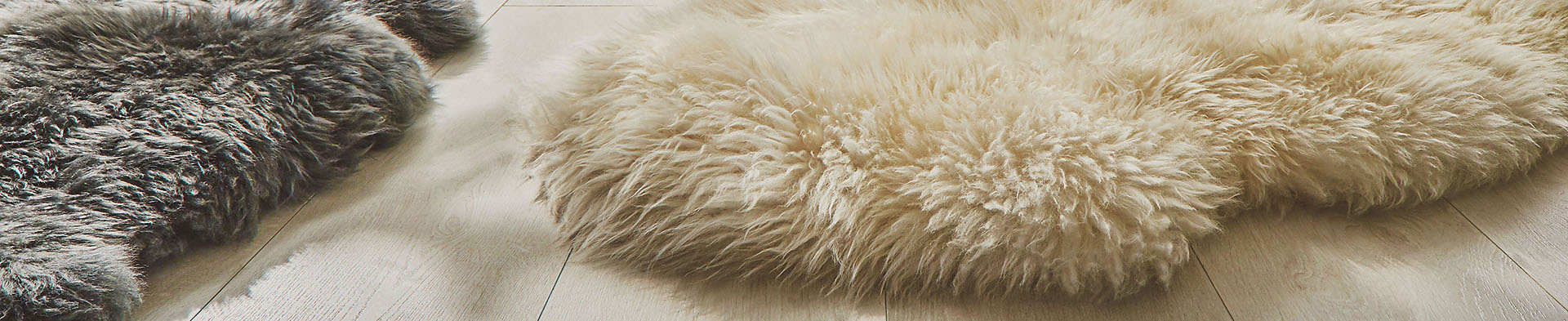 Zwei Fellteppiche liegen nebeneinander auf hellem Holzboden, einer grau, einer beige.
