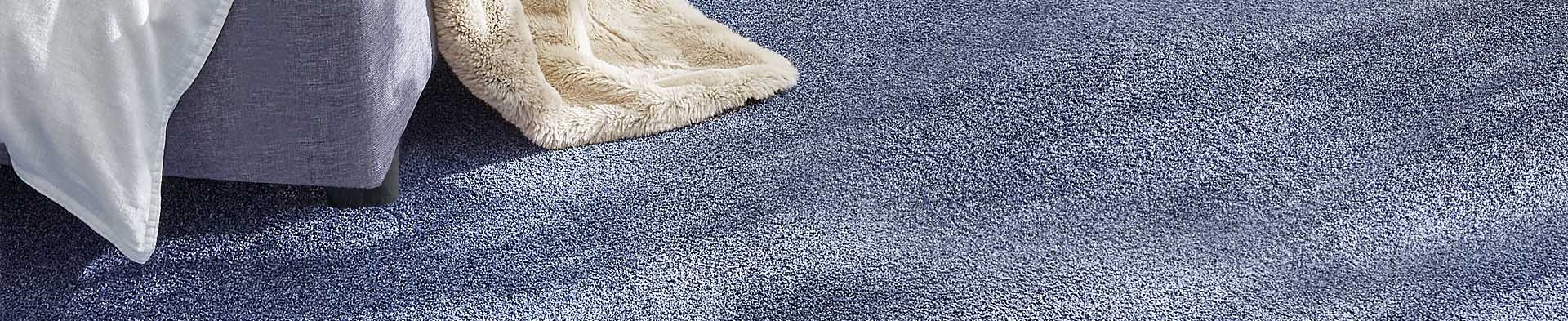 Bettkante mit weißer Decke und beiger Decke auf blauem Teppichboden in Innenraum.