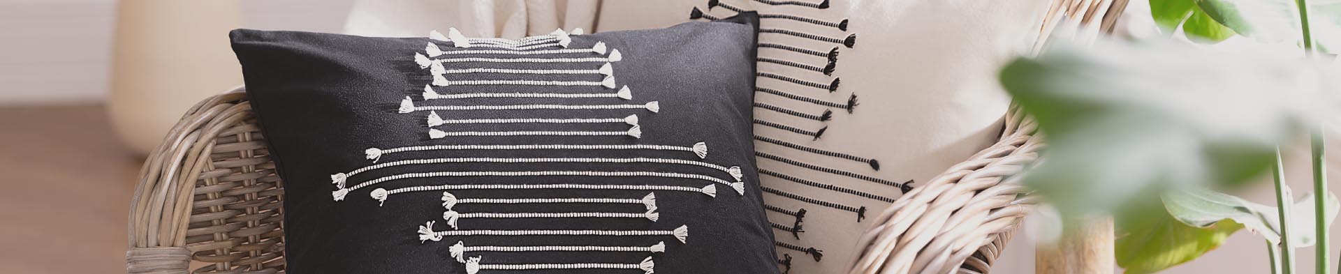 Zwei Kissen mit gestreiften Mustern liegen auf einem Korbstuhl neben grünen Blättern.