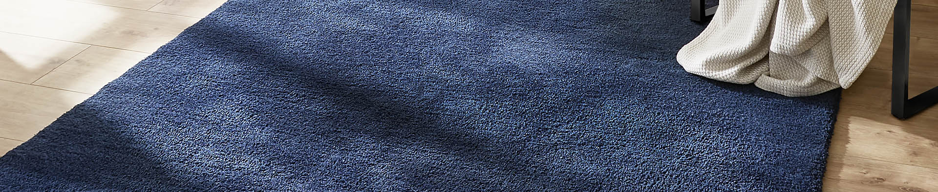 Blauer Teppich liegt auf Holzboden, eine weiße Decke hängt über Stuhlbein im hellen Raum.