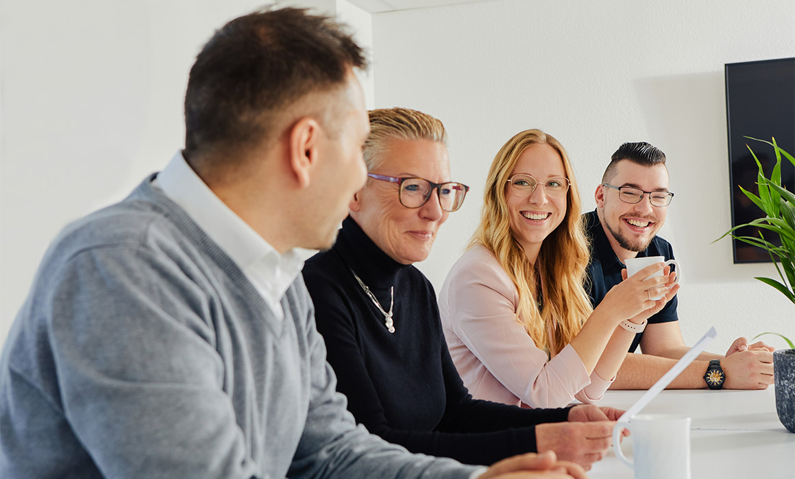 Vier Personen sitzen an einem Tisch, unterhalten sich und trinken Kaffee in einem Büro.