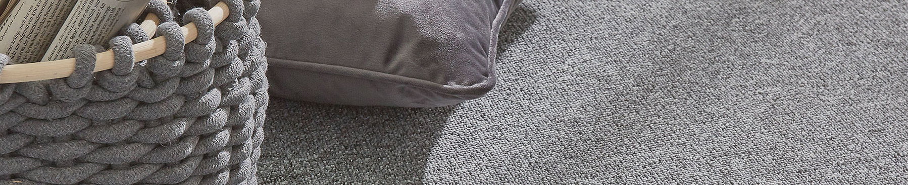 Geknüpfter Korb hält Zeitung, liegt neben einem Kissen auf grauem Teppichboden.