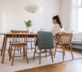 Frau sitzt am Holztisch und liest in einem hellen, minimalistischen Esszimmer mit Fenstern.