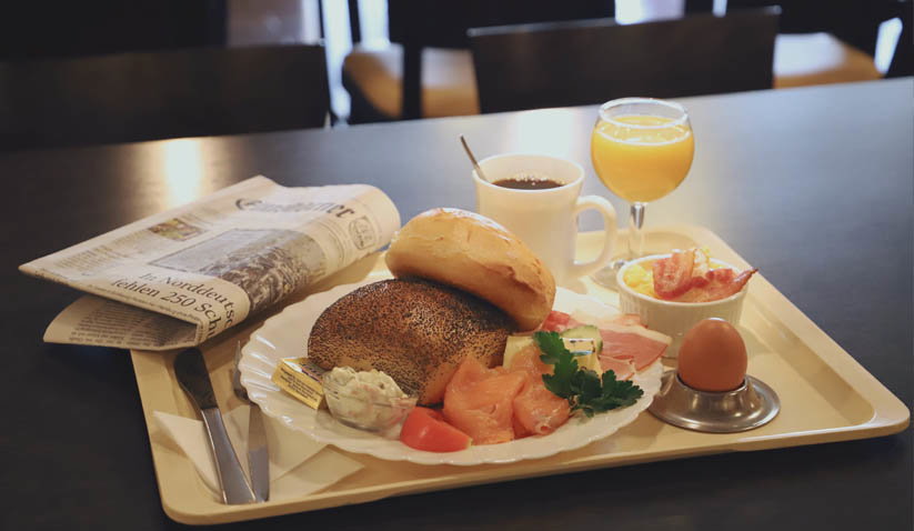 Frühstückstablett mit Brötchen, Lachs, Butter, gekochtem Ei, Kaffee, Orangensaft und Zeitung auf Tisch.