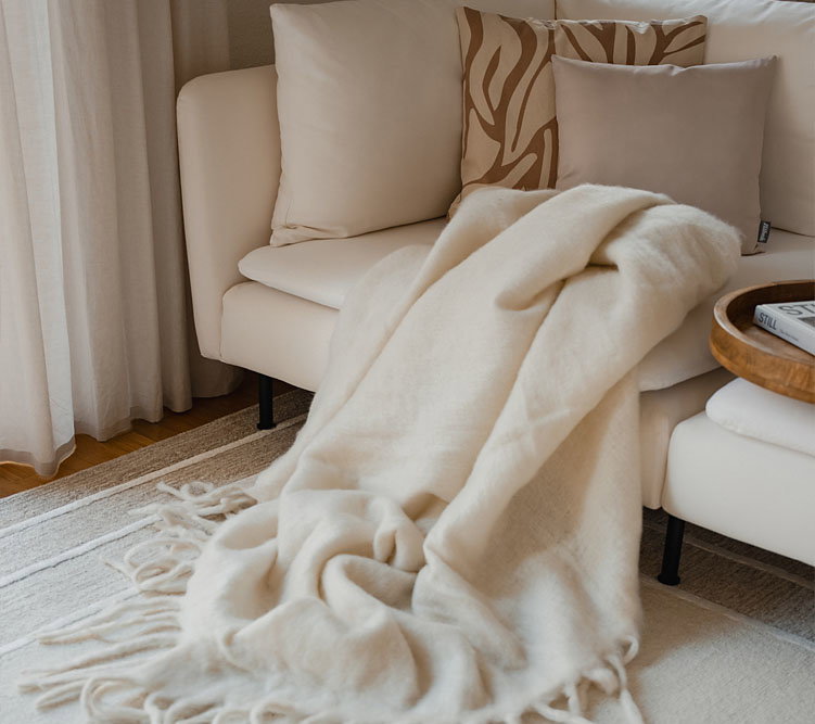 Beige Decke liegt auf einem cremefarbenen Sofa mit Kissen in einem hellen Wohnzimmer.