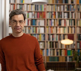 Mann steht vor Bücherregal, trägt rotbraunen Pullover, Bücherwand unscharf im Hintergrund.