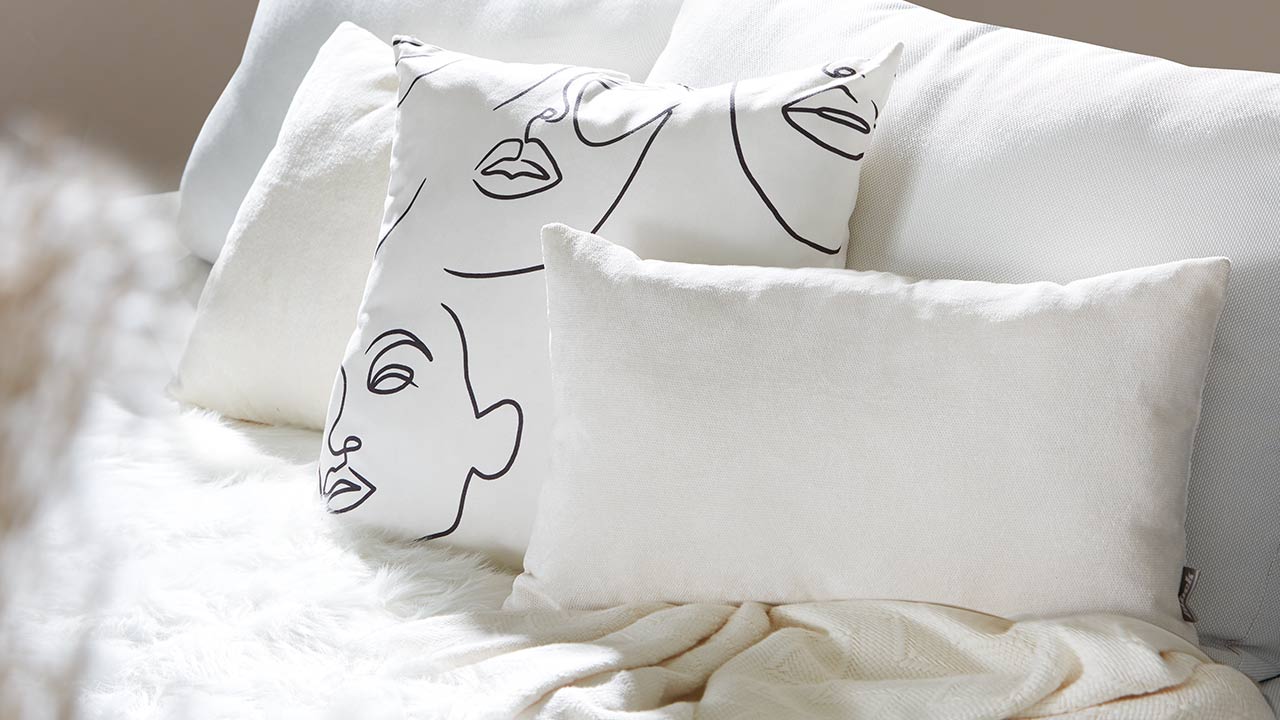 Weiße Kissen, eins mit abstraktem Gesichtsmuster, liegen gemütlich auf einem Sofa mit Decke.