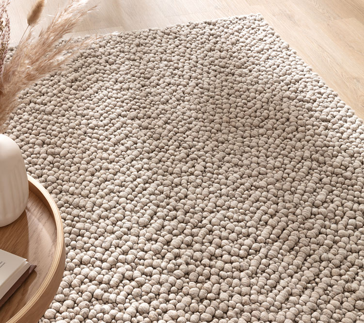 Beigefarbener Teppich mit dicker, gepunkteter Textur liegt auf hellem Holzboden neben Holztablett mit Vase.