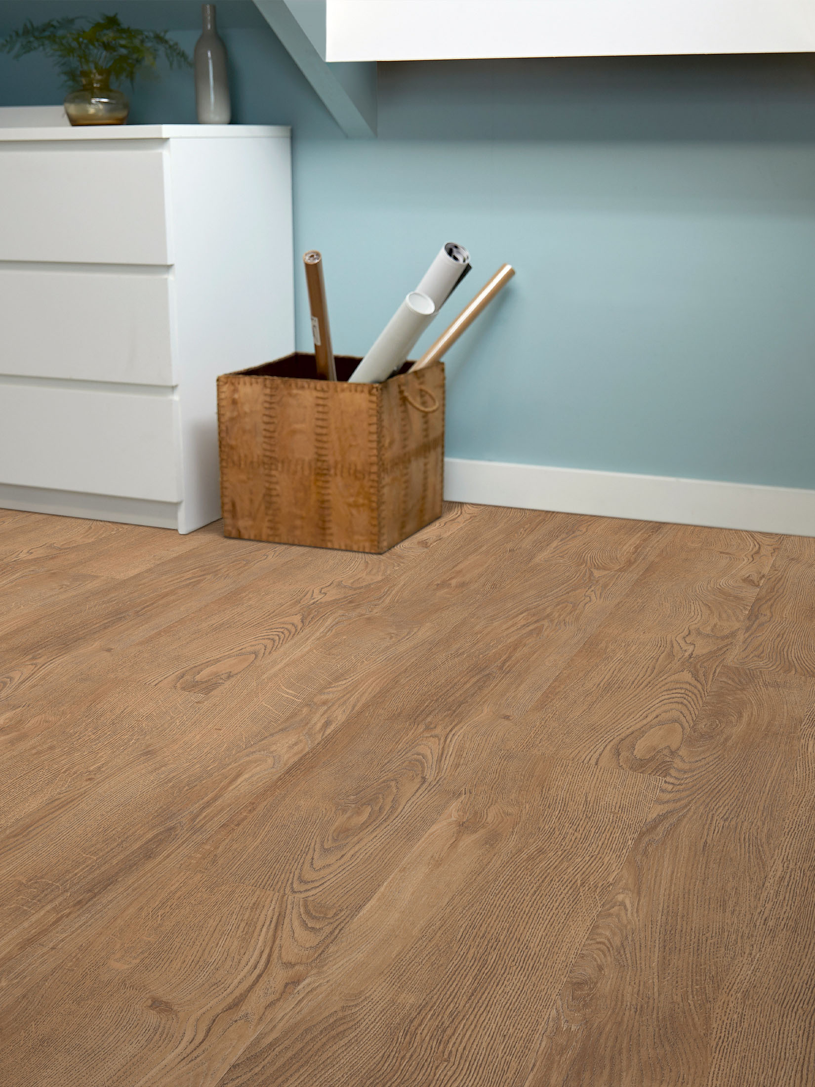 Holzkorb mit gerollten Papieren steht auf Holzfußboden neben weißem Schrank vor blauer Wand.
