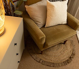 Grüner Sessel steht auf rundem Juteteppich neben weißem Schrank mit Lampe und Pflanzen.