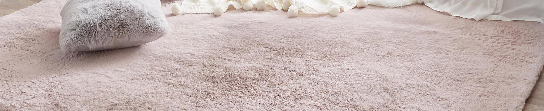 Rechteckiger, rosa Teppich liegt auf hellbraunem Holzboden, graues Kissen und weiße Decke am Rand.