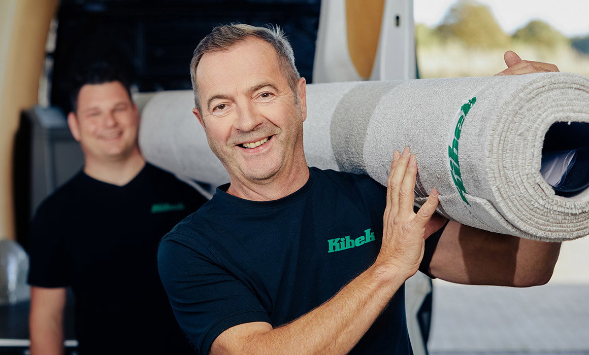 Mann trägt Teppichrolle auf Schulter, zweiter Mann steht vor Lieferwagen, beide tragen Kibek-Shirt.