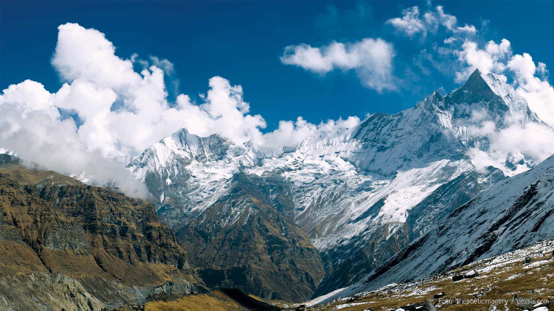 Schneebedeckte Berge mit Wolken am klaren blauen Himmel über trockenem Tal im Vordergrund.
