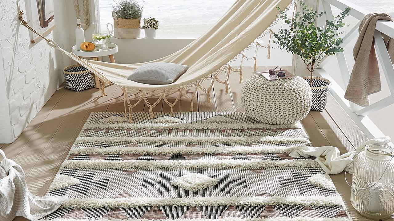 Hängematte hängt mit Kissen auf Holzterrasse mit Teppich, Pflanzen und Pouf am Fenster.
