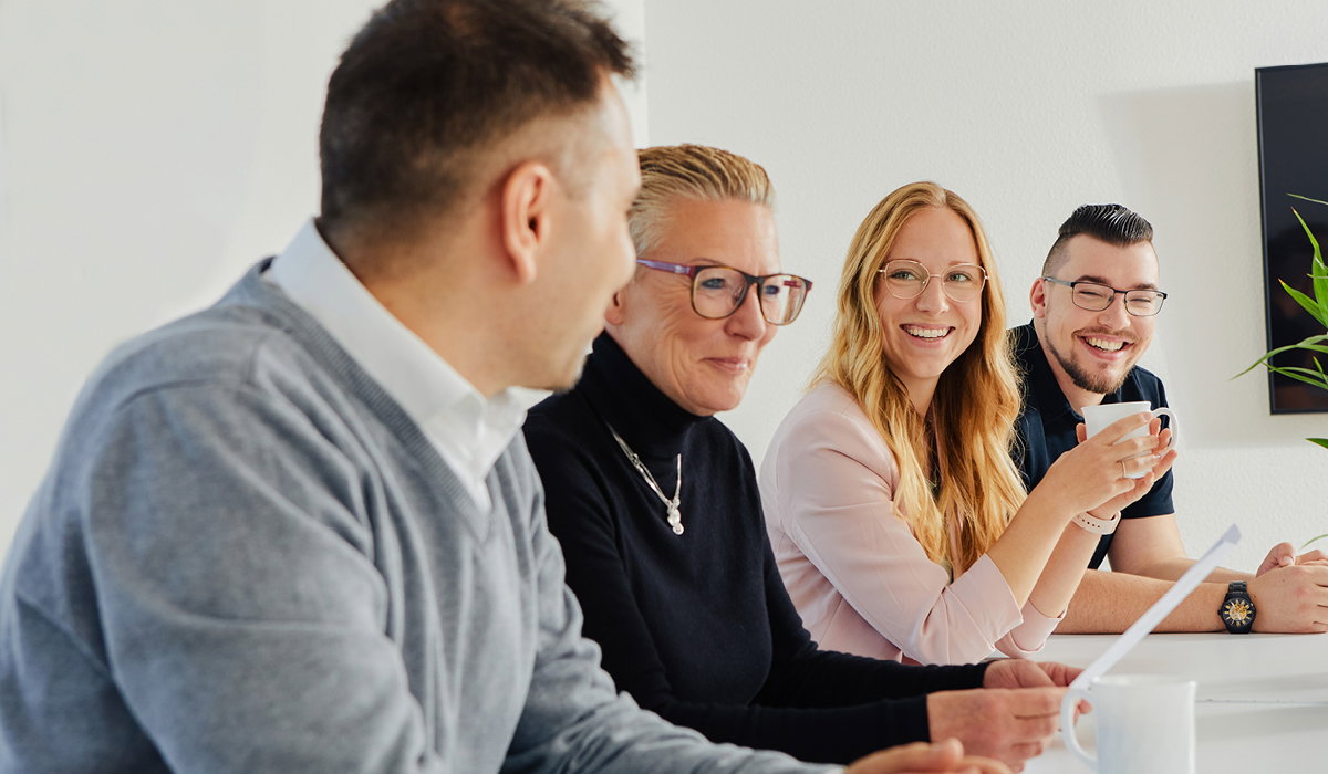 Vier Personen unterhalten sich lächelnd bei einem Treffen in einem hellen Büro.