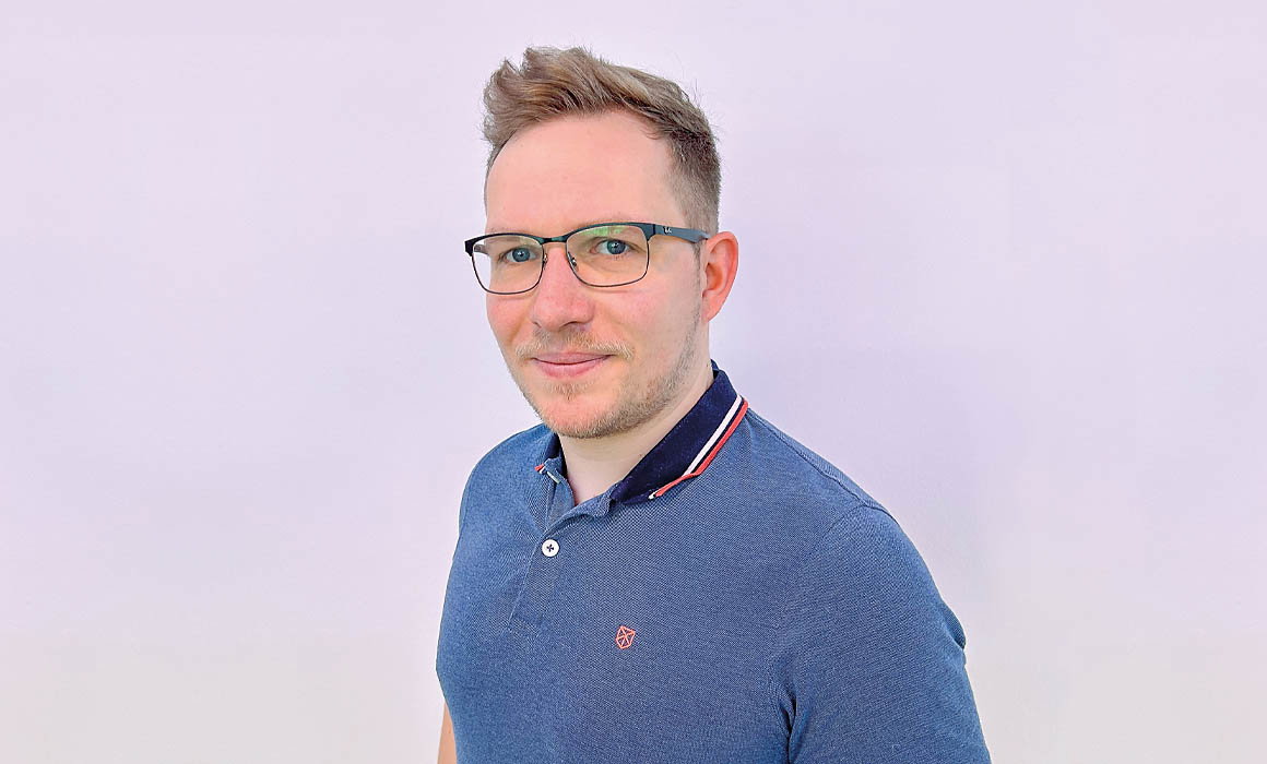 Mann mit Brille trägt blaues Polohemd und steht vor weißem Hintergrund.