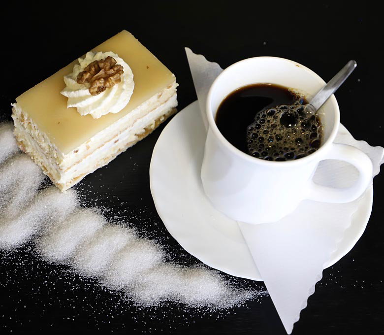 Kaffeetasse mit Löffel auf weißem Unterteller, daneben ein Stück Kuchen mit Walnuss, Zuckerstreifen.