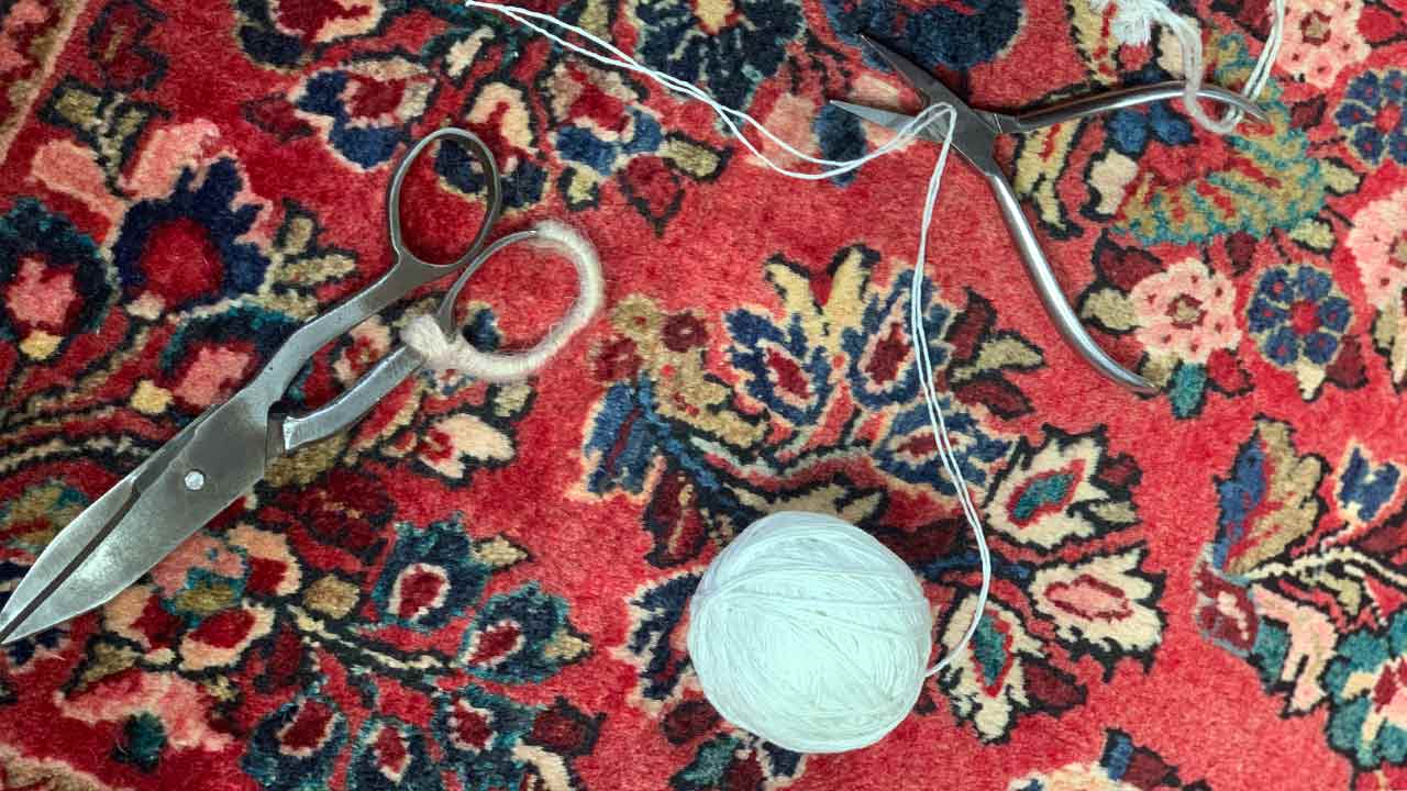 Schere und Zange liegen neben einem weißen Wollknäuel auf einem gemusterten roten Teppich.