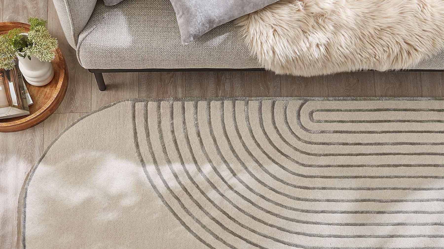 Beiger Teppich mit grauen Linienmuster liegt auf Holzboden vor grauem Sofa mit Felldecke.