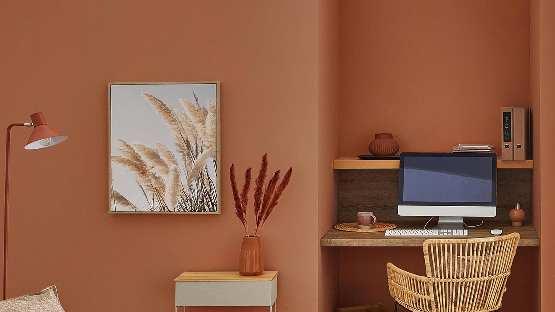Schreibtisch mit Computer und Stuhl, daneben Vase mit roten Zweigen, dekorativer Raum mit Bild und Lampe.