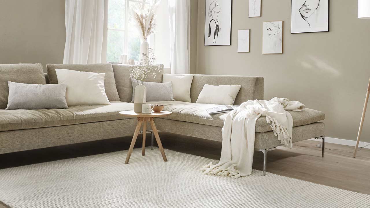 Ecksofa mit Kissen und Decke vor Fenster, kleiner Tisch mit Vase im hellen Wohnzimmer.