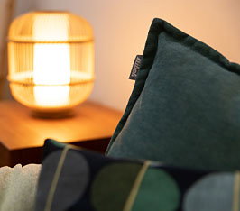 Kissen liegen auf einem Sofa, im Hintergrund leuchtet eine Tischlampe auf einem Holztisch.