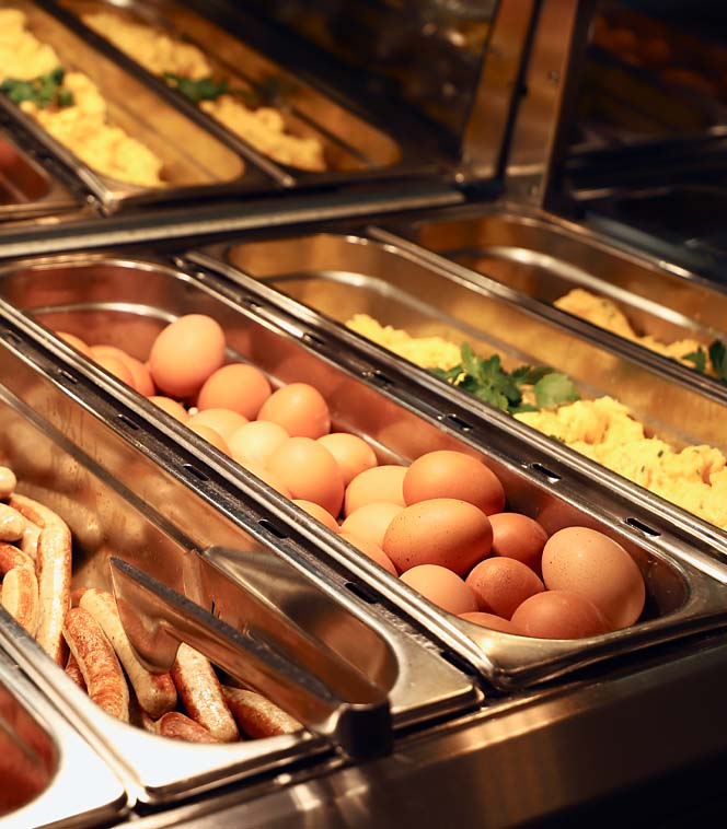 Eier, Würstchen und Rührei liegen in Metallbehältern auf einem Buffet unter Wärmelampen.