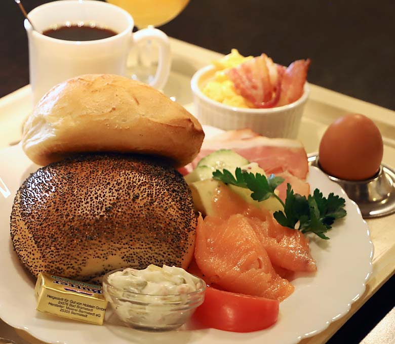 Brötchen liegen neben Lachs, Ei, Bacon, Salat und Butter auf Teller vor Kaffee und Orangensaft.
