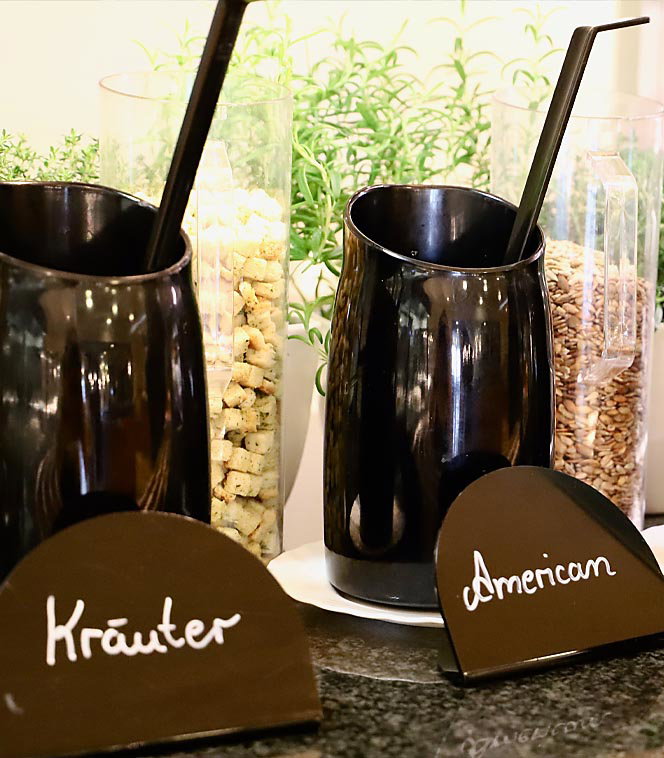 Zwei schwarze Becher mit Löffeln stehen vor Glasbehältern, Beschriftung Kräuter und American.