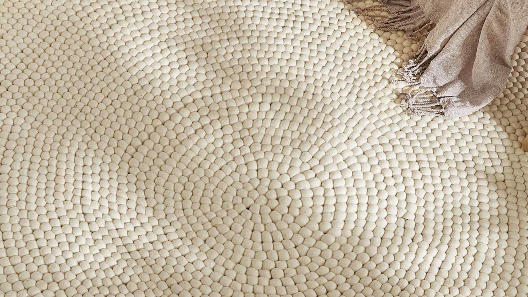Runder, beiger Teppich mit gestrickter Struktur liegt auf dem Boden, oben rechts Decke.
