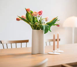 Blumenstrauß steht in Vase auf Holztisch, daneben zwei brennende Kerzen, Stühle im Raum.