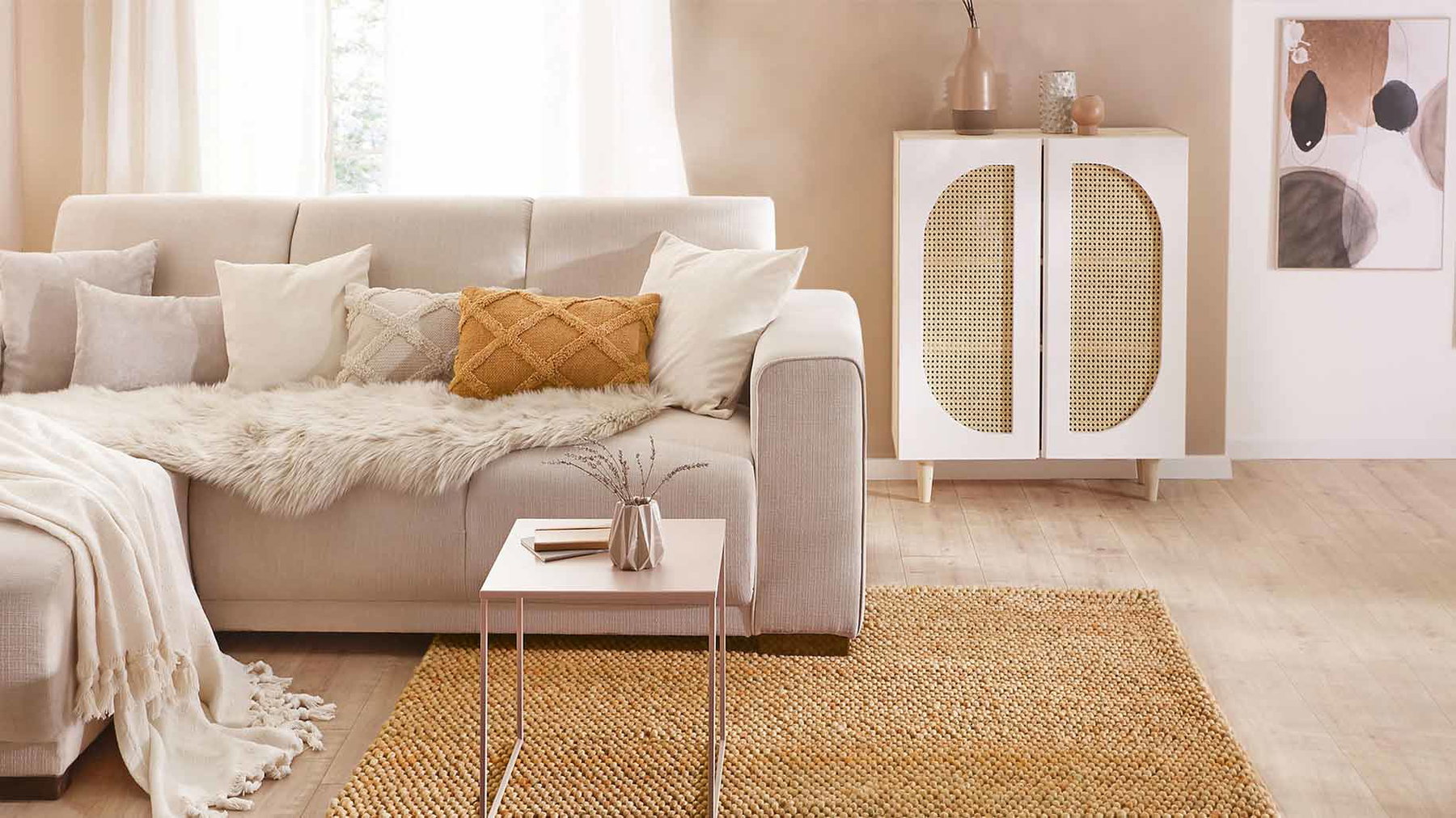 Beige Sofa mit Kissen und Decke steht neben kleinem Tisch und kleinem Schrank in hellem Raum.