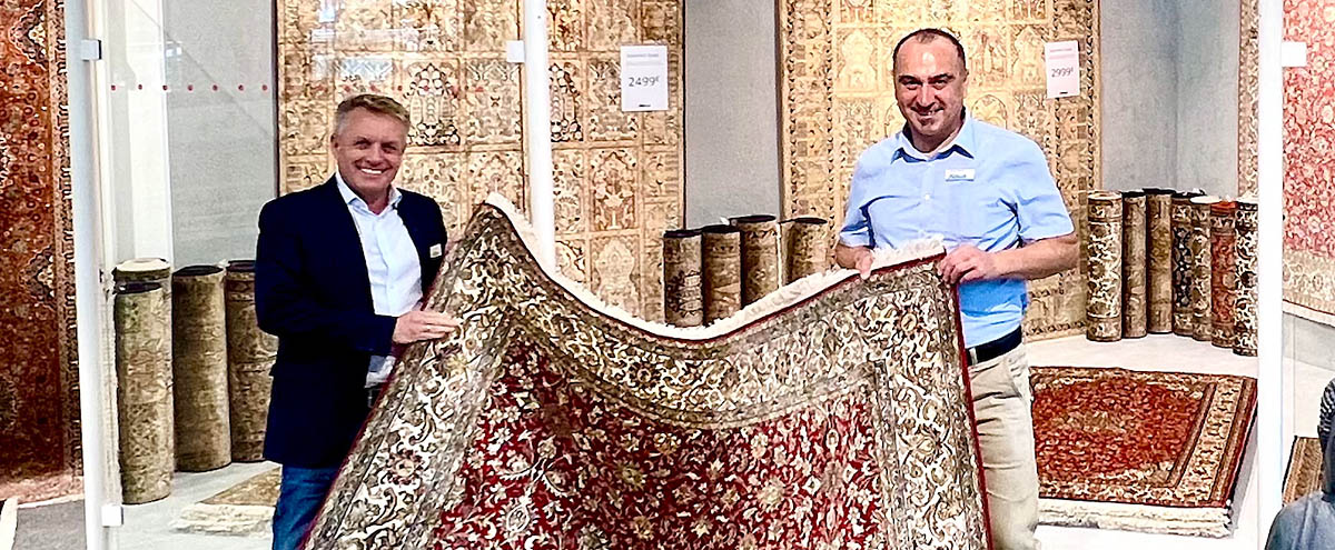 Zwei Männer halten einen Teppich mit floralem Muster in einem Teppichgeschäft mit Rollen im Hintergrund.