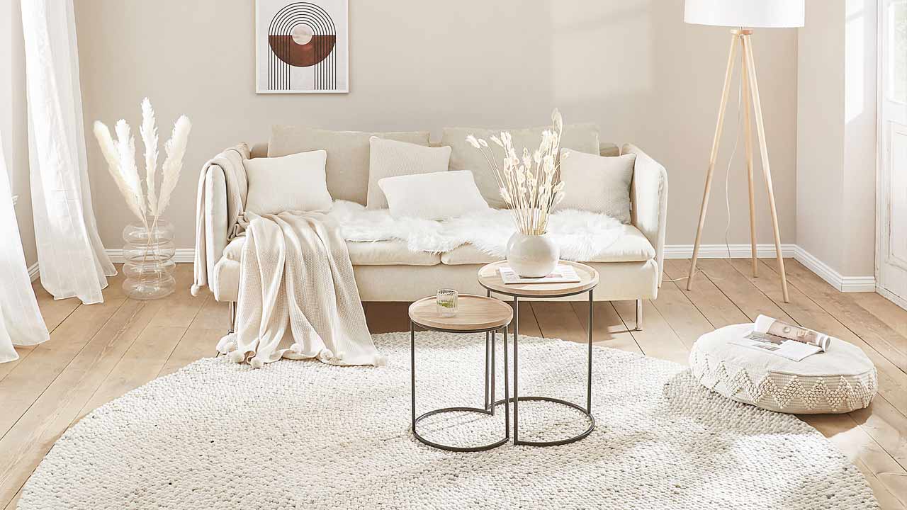 Beiges Sofa mit Kissen und Decke steht im hellen Wohnzimmer mit Teppich, Tisch und Stehlampe.