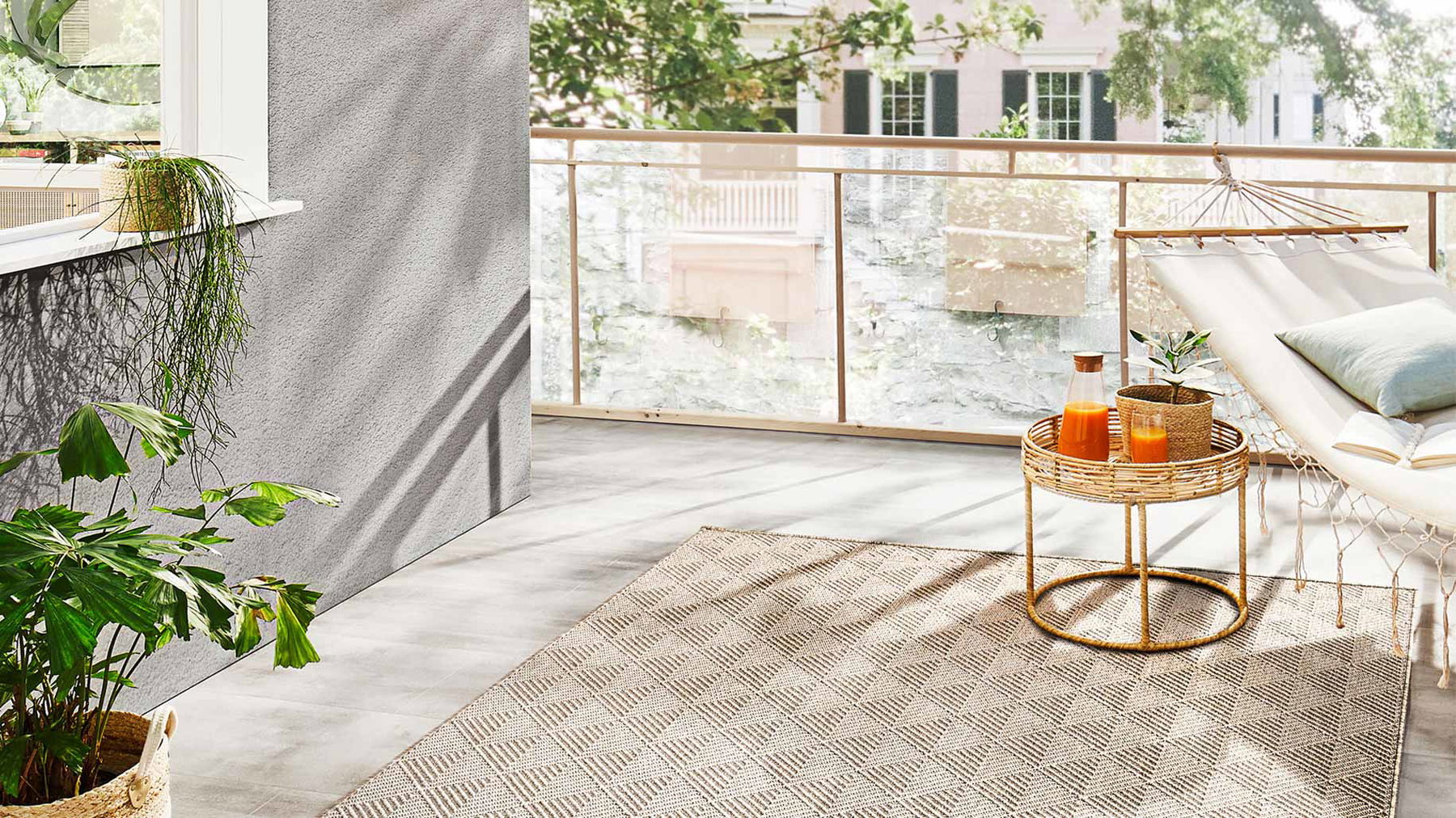 Terrassenmöbel mit einer Hängematte und einem kleinen runden Tisch mit Saft und Pflanze auf einem Teppich, Sonnenlicht draußen.