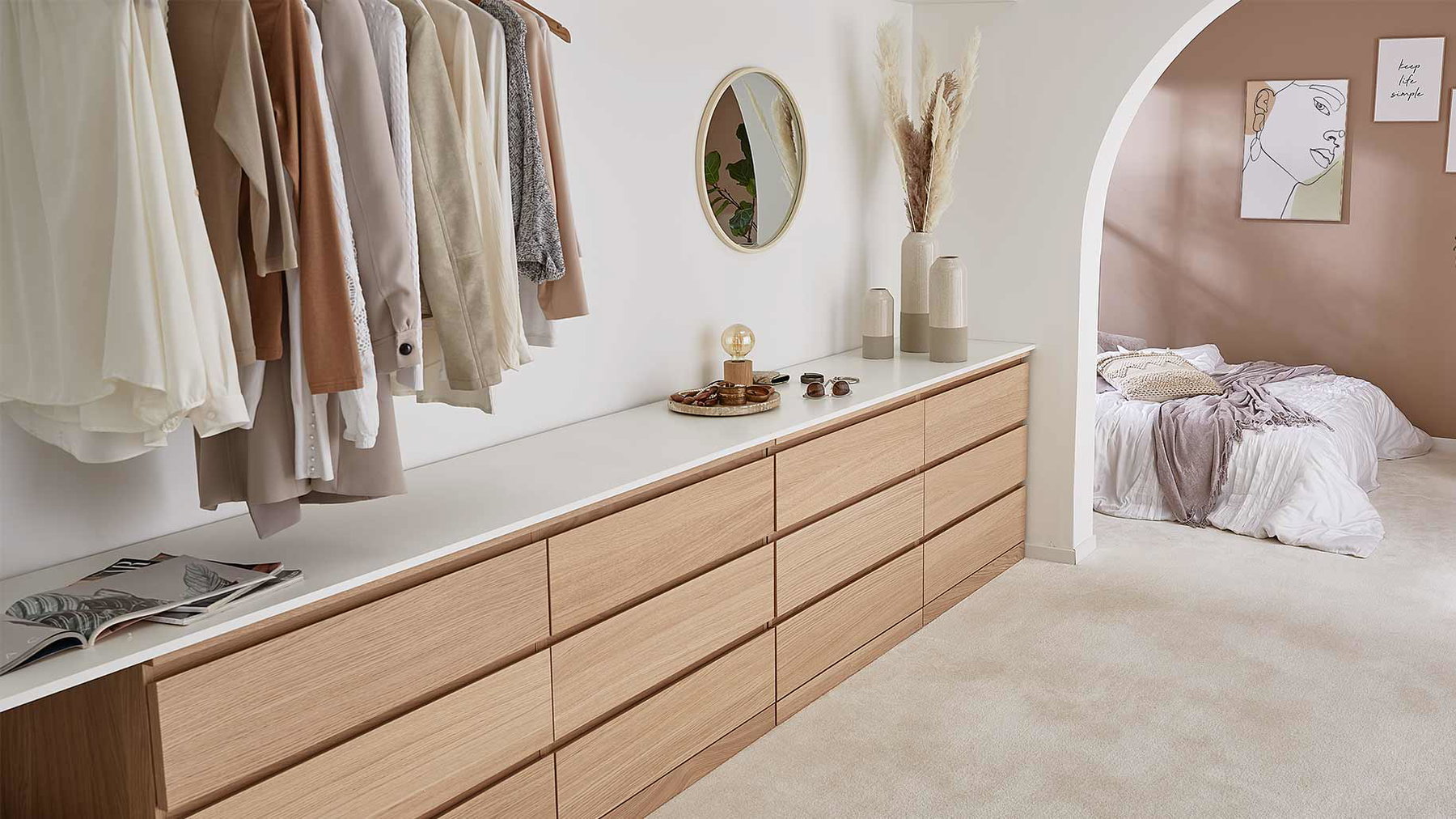 Sideboard mit Kleidung hängend darüber, Spiegel und Vasen, daneben Bett durch Bogenrahmen sichtbar.