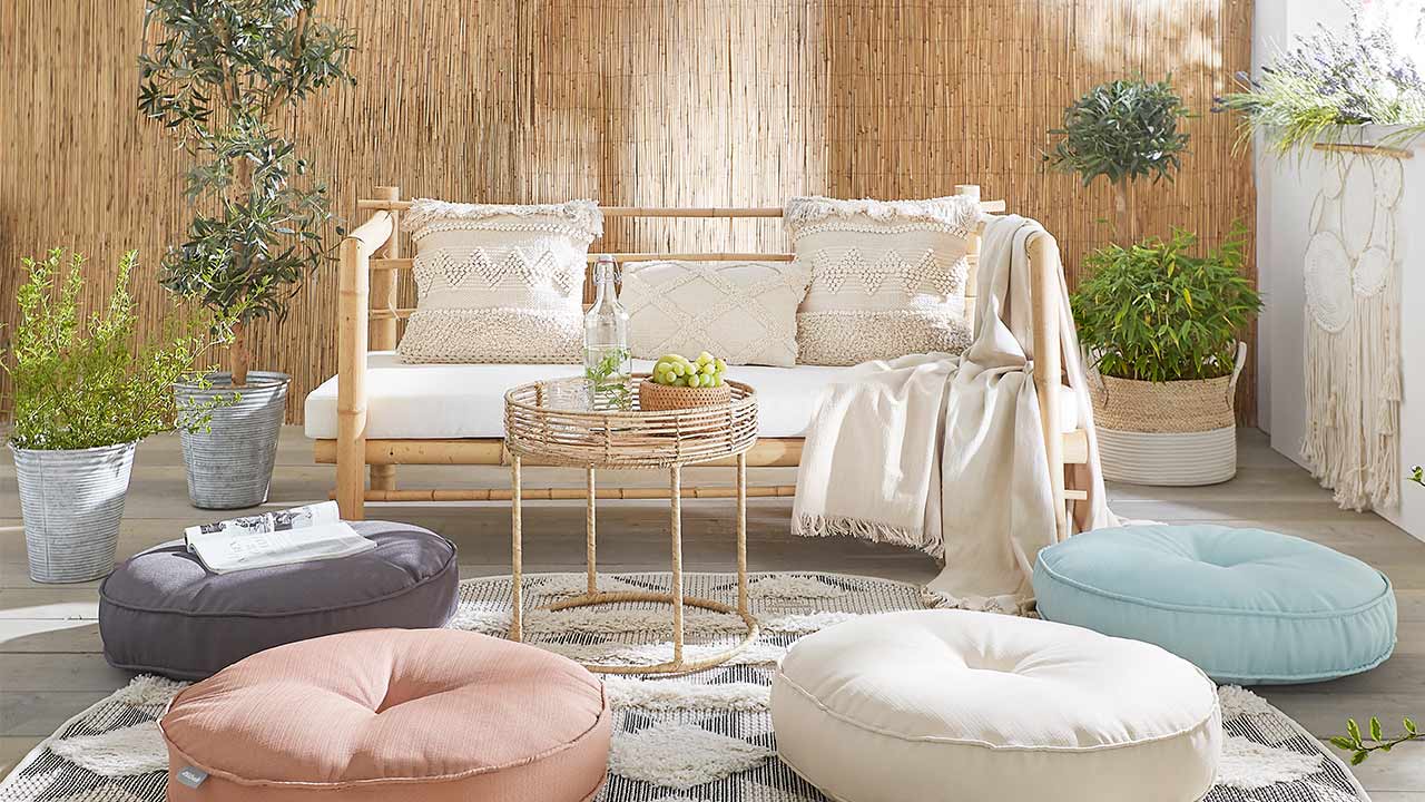 Sofa mit Kissen und Decke, runder Tisch mit Flasche und Früchten, Bodenkissen und Pflanzen im Raum.