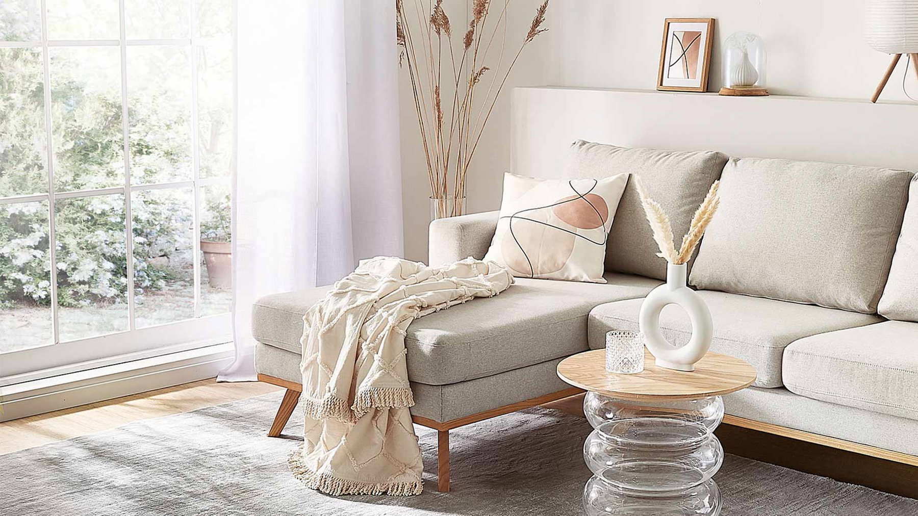 Beige Ecksofa mit Decke und Kissen steht im hellen Wohnzimmer mit Fenster und Tisch.