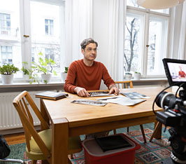 Mann sitzt am Tisch und blättert in Zeitungen, helle Wohnung mit Fenstern im Hintergrund.