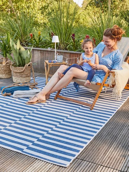 Frau sitzt mit Kind auf Liegestuhl auf Terrasse Teppich mit Streifen Pflanzen im Hintergrund