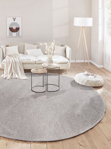 Runder grauer Teppich liegt im Wohnzimmer vor Sofa mit Kissen und zwei Beistelltischen.