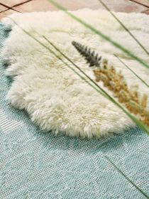 Weiße flauschige Unterlage liegt auf hellblauem Teppich mit Gräsern im Vordergrund