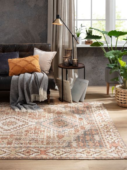 Brauner Teppich mit geometrischem Muster liegt auf Holzboden vor schwarzem Sofa mit Kissen und Decke