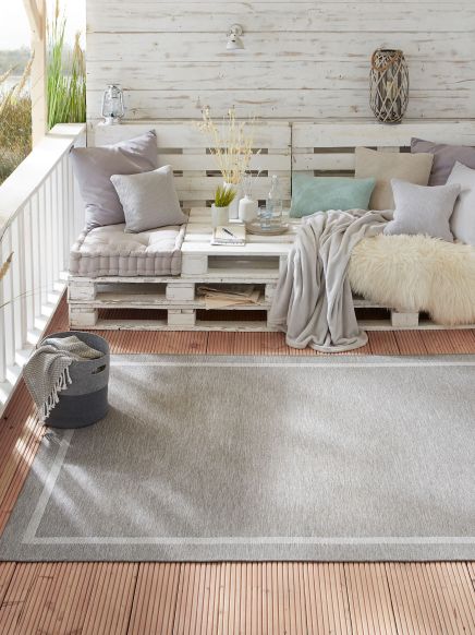 Sitzbank mit Kissen und Decken auf Holzterrasse, Teppich und Pflanzen im Außenbereich.